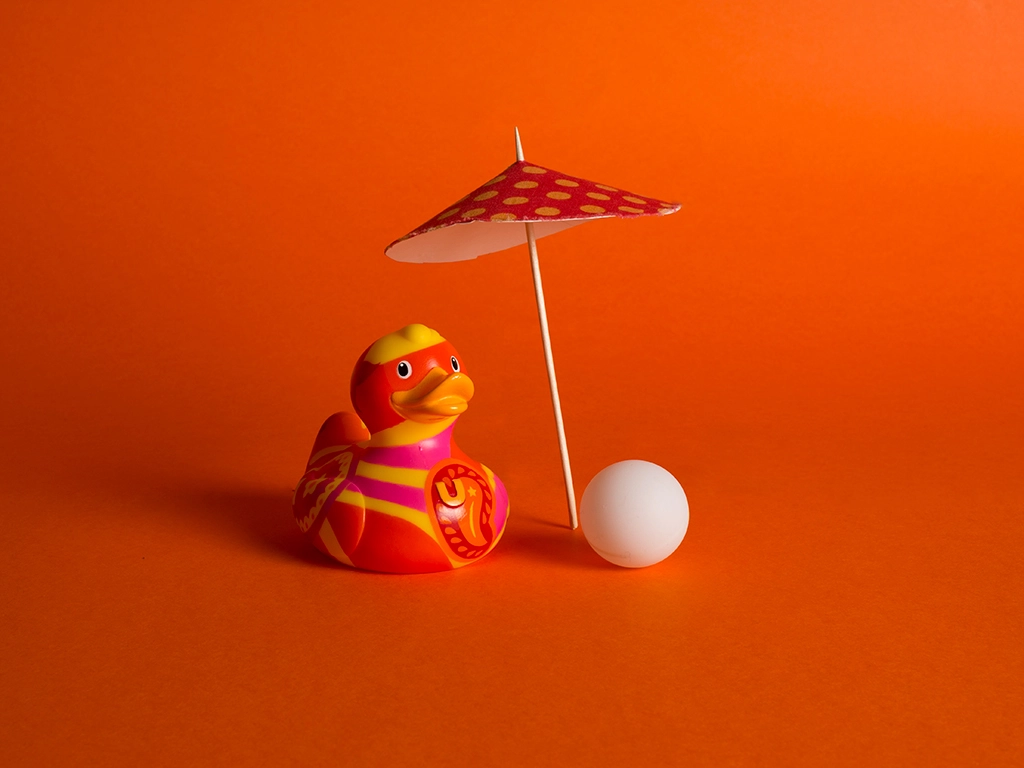 Canard en plastique à motif aléatoire orange et rouge avec un parasol à pois dans les même tons et une balle blanche sur fond orange