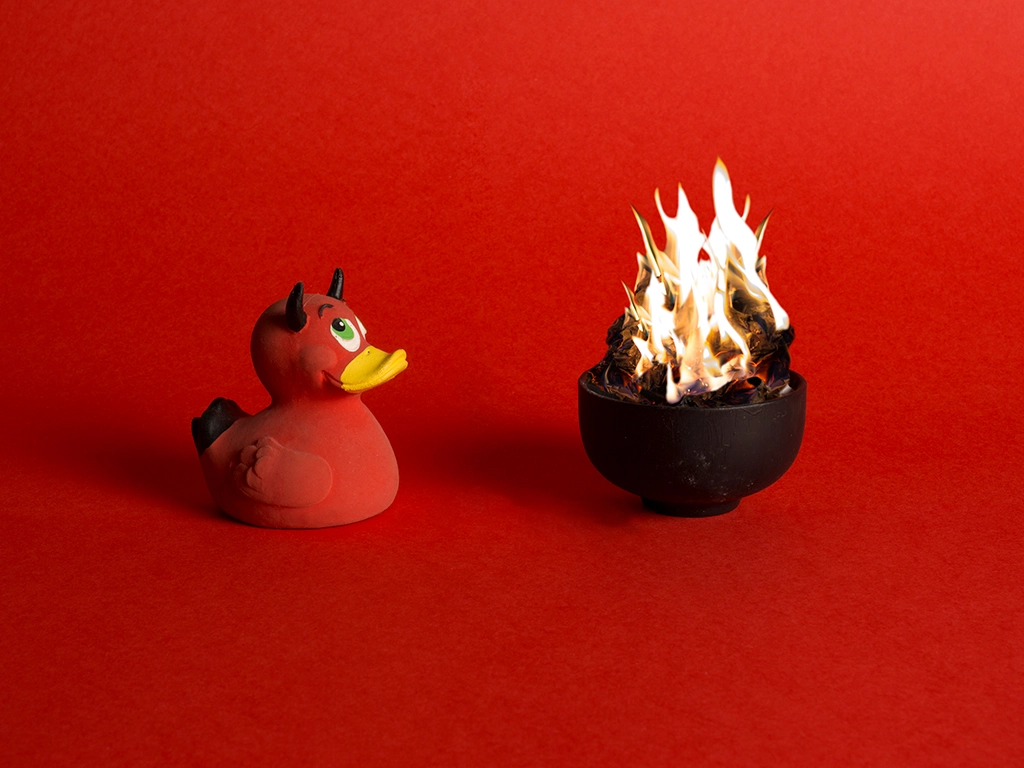 Canard en plastique rouge avec des petites cornes de diable regardant un feu brûler dans une petit pot noir sur fond rouge