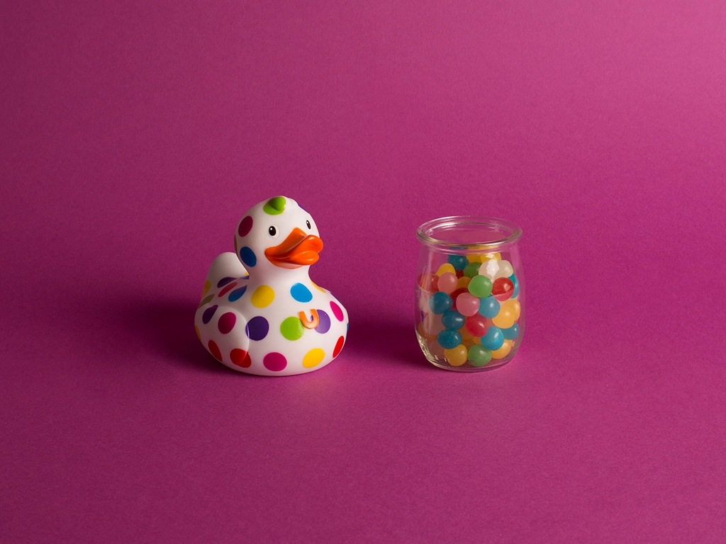 Canard en plastique blanc avec des pois de couleur et un petit pot en verre rempli de bonbons à ses côtés sur un fond violet